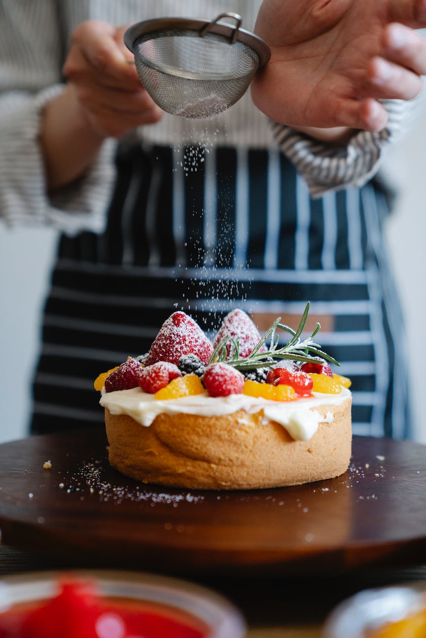 man in a striped apron sprinkling powdered sugar on a cake with strawberries